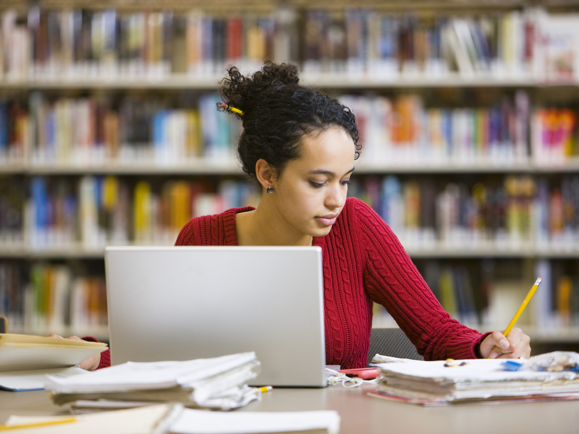 Black female college student sitting at a desk in a library on campus. She has her laptop open, and is writing notes in a notebook with a pencil.